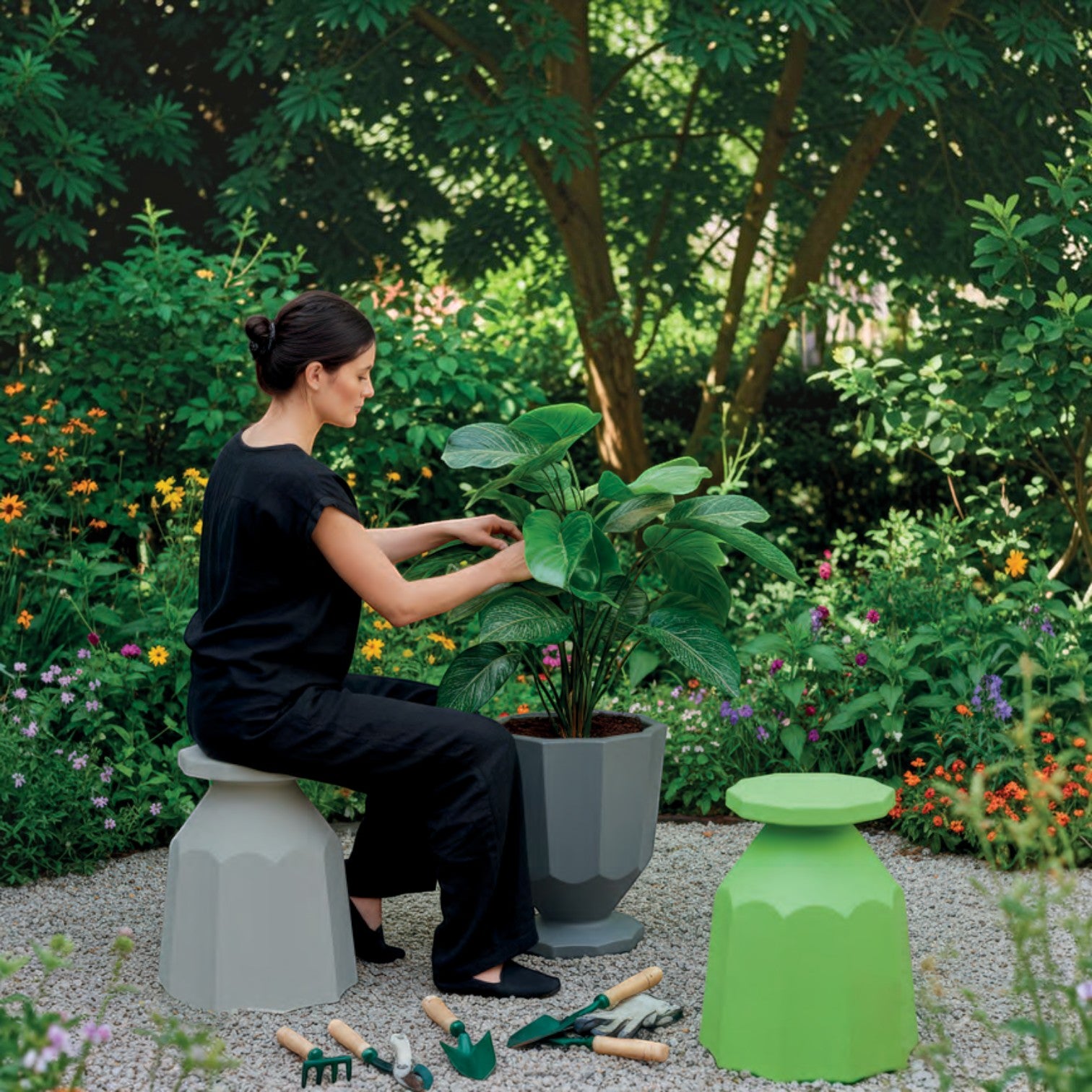 Addo Shop Caimi recycled grey planter with a large green plant in a garden setting with a woman sitting on a stool.