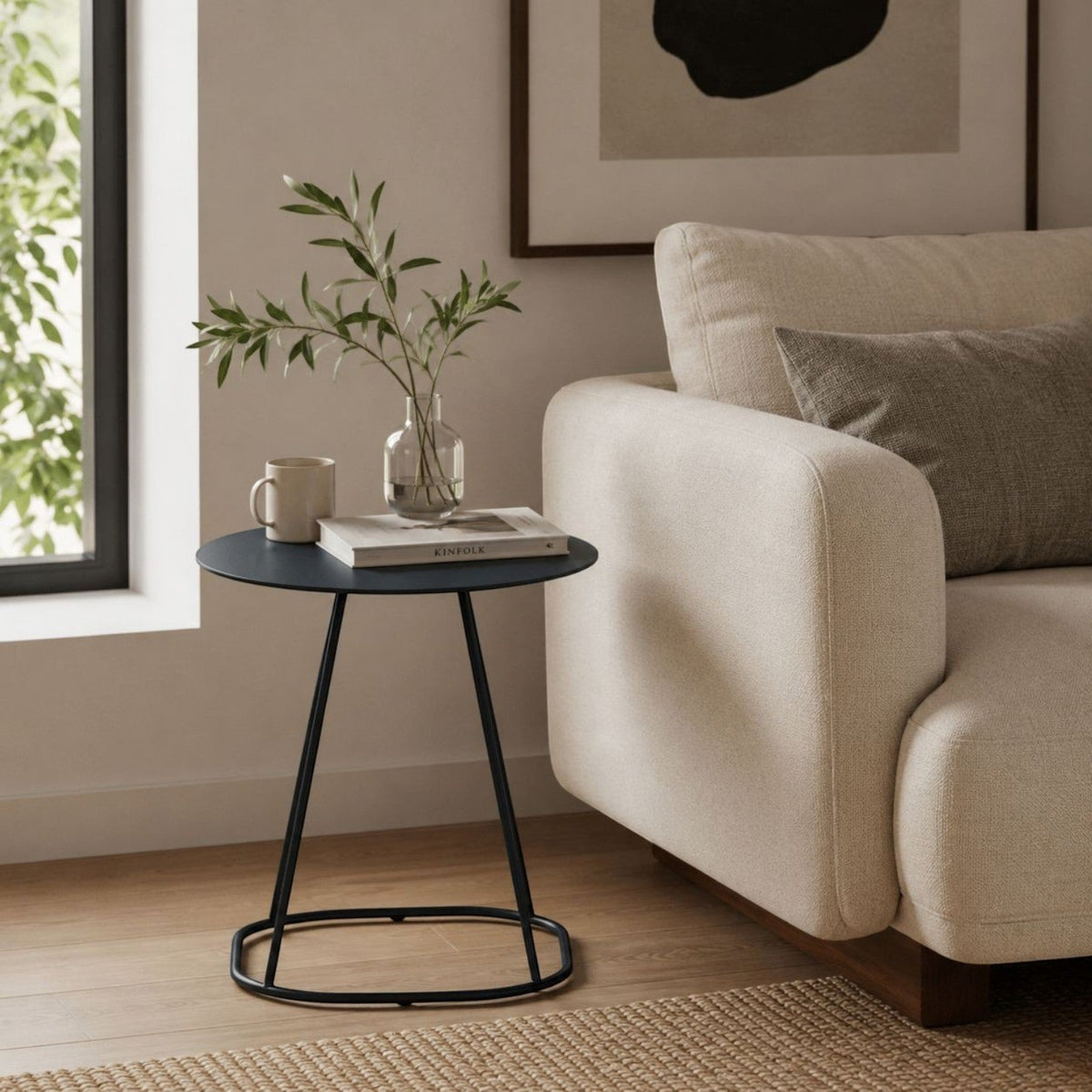 Addo Shop Kiran round side table with a matte black metal base and tabletop next to a beige sofa in a living room.