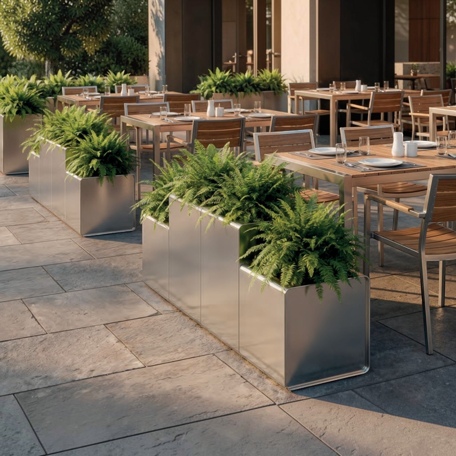 Addo Shop PRISMA outdoor planter in brushed metal with a tiered design, filled with lush green ferns on a patio.
