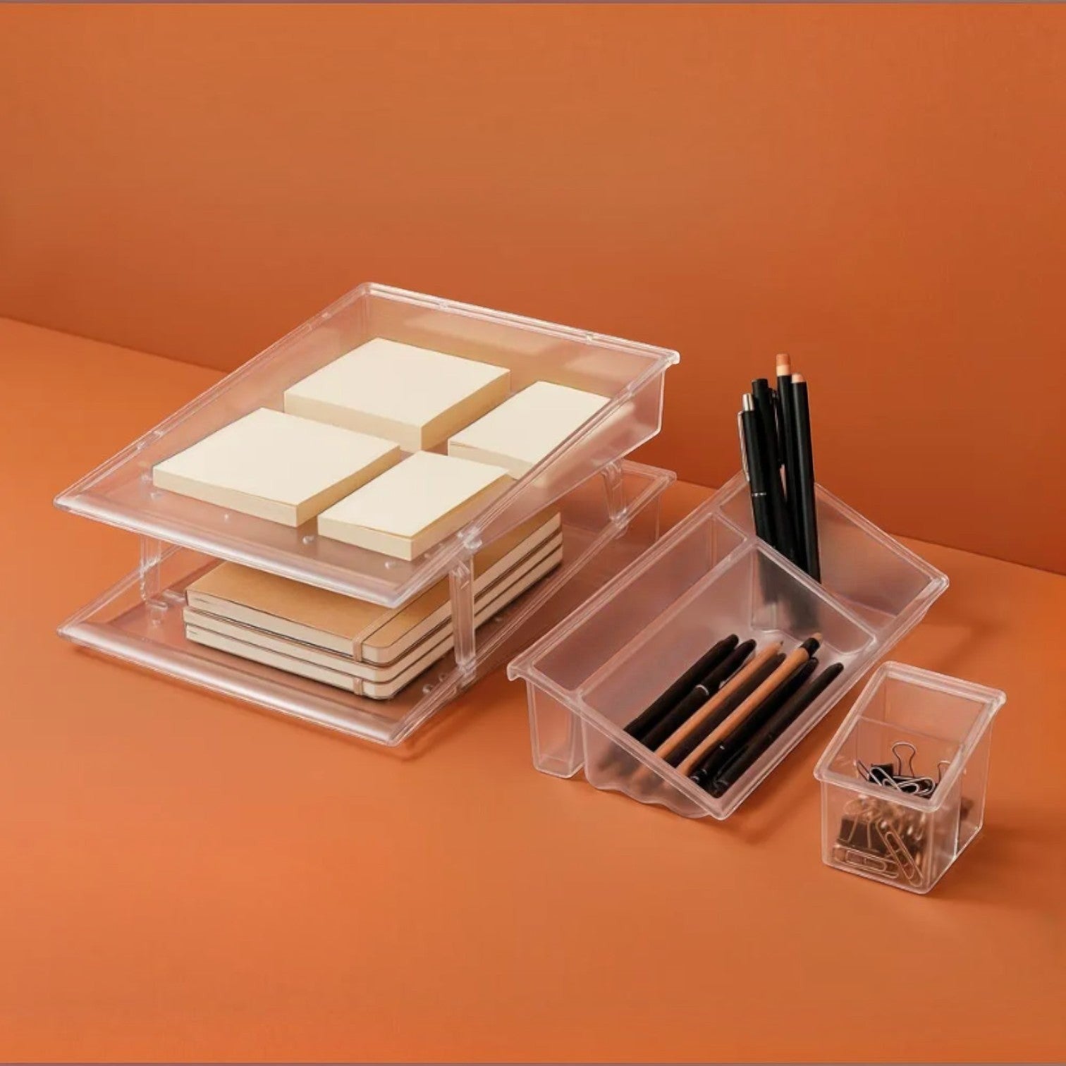 Addo Shop clear plastic stacking desk organizers holding notebooks, sticky notes, pens, and binder clips on orange.