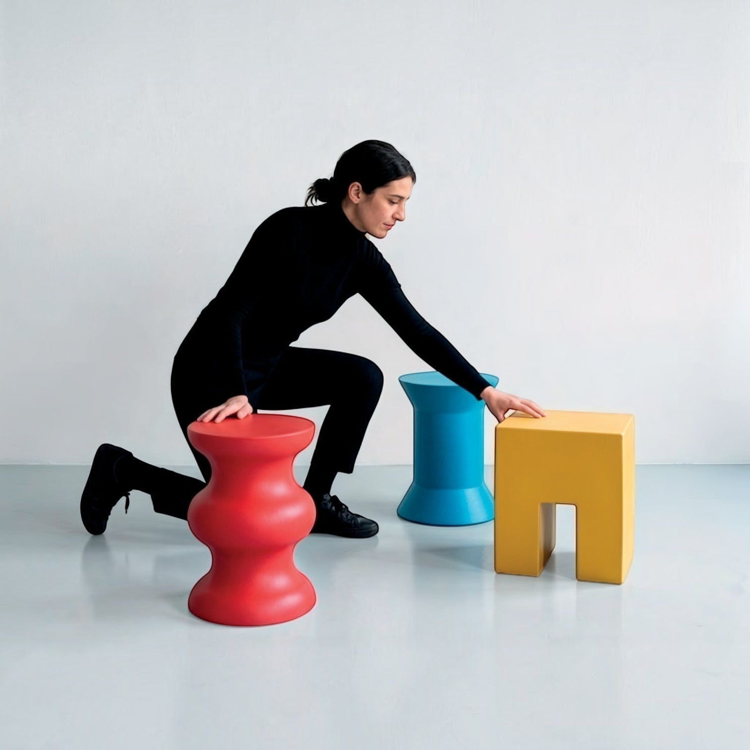Woman in black kneeling next to a yellow STILEMI Cube stool by Addo Shop, with red and blue stools nearby.